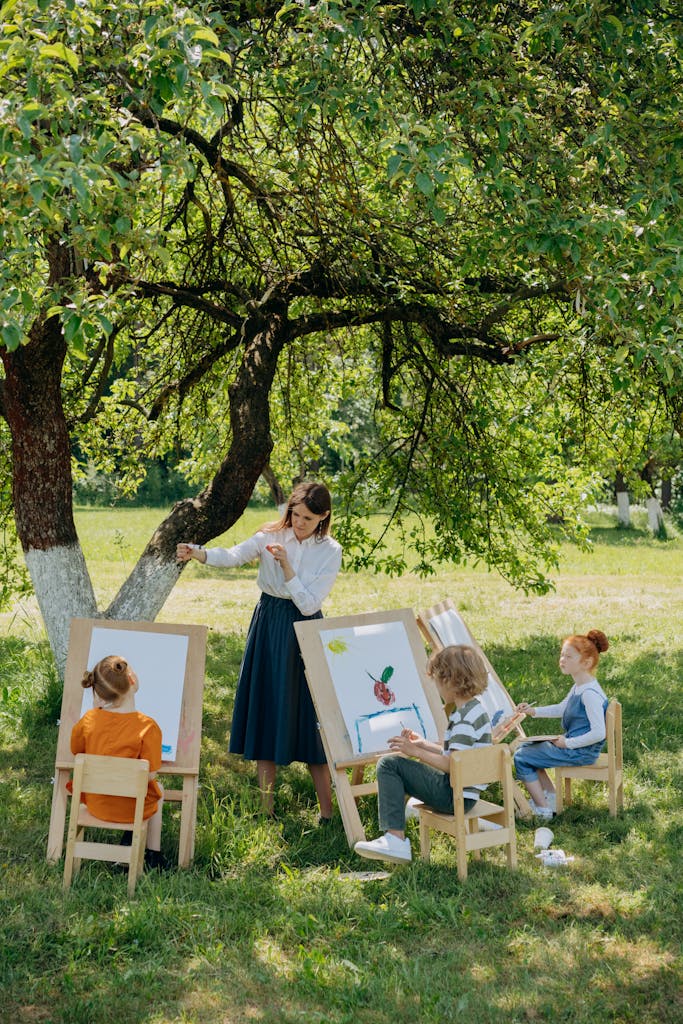 Children enjoying painting outdoors under a tree guided by a teacher on a sunny day.