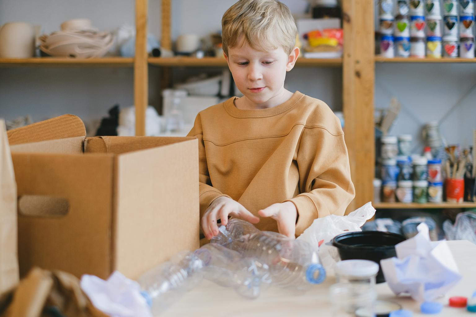 Young boy exploring plastic recycling indoors with cardboard boxes and bottles around.